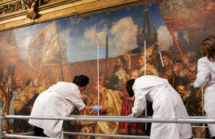 Zdjęcie przedstawia trzy konserwatorki w białych fartuchach pracujące przy renowacji obrazu.